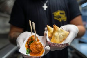 A person holding two dishes as if offering them to the viewer. The first dish is something like a meatball and the second is two samosas.